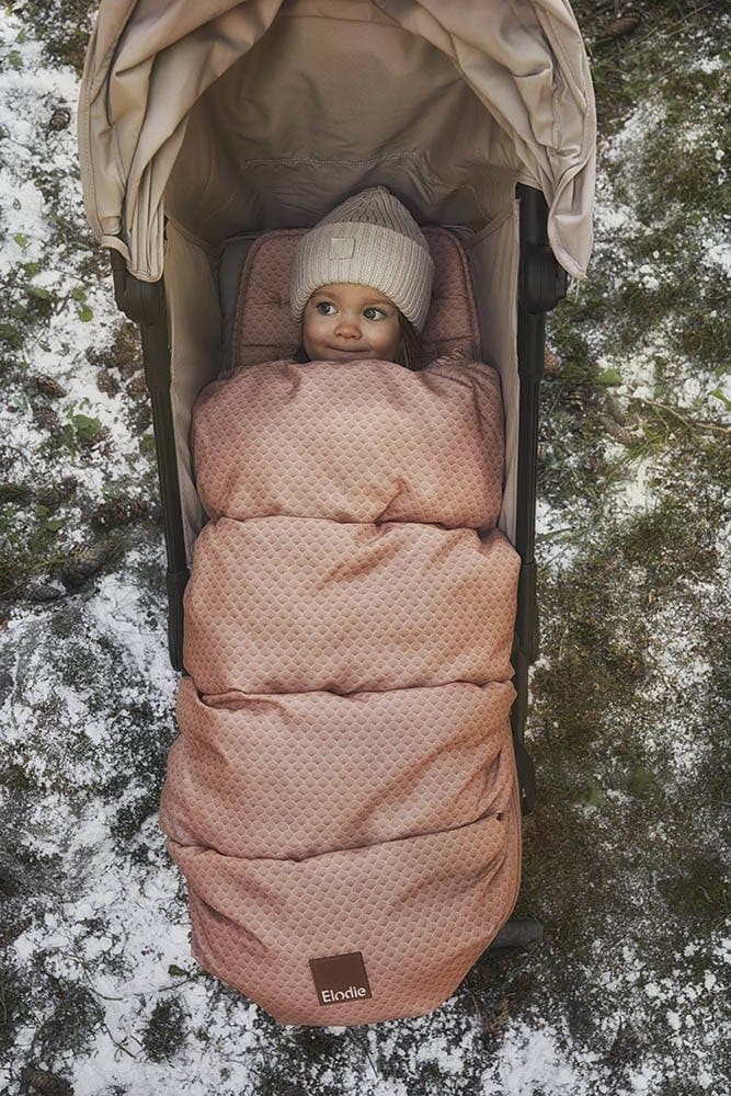 Różowy śpiworek i wkładka do wózka z delikatnym wzorem dla dziecka w zimowej czapce w wózku na tle śniegu i trawy. Elodie Details śpiworek i wkładka do wózka 2w1 Pink Noveau