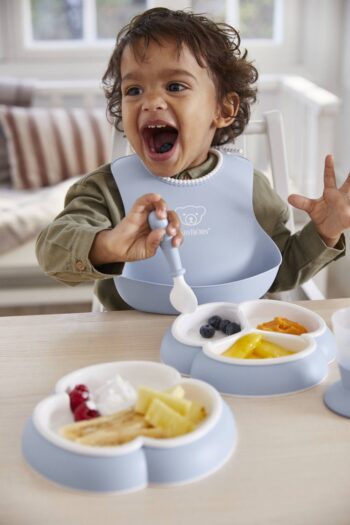 Dziecko z niebieskim silikonowym śliniakiem i niebieskim zestawem obiadowym z podziałem na trzy części z jedzeniem dla dziecka. BABYBJORN Zestaw obiadowy Dinnerware, 8 szt. Powder Blue