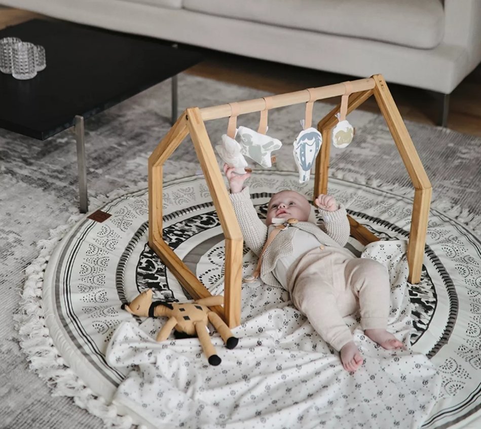 Drewniany stojak edukacyjny w kształcie domku z zawieszonymi zabawkami dla niemowlaka leżącego na okrągłym dywanie. Elodie Details House of Elodie Baby Gym Stojak edukacyjny