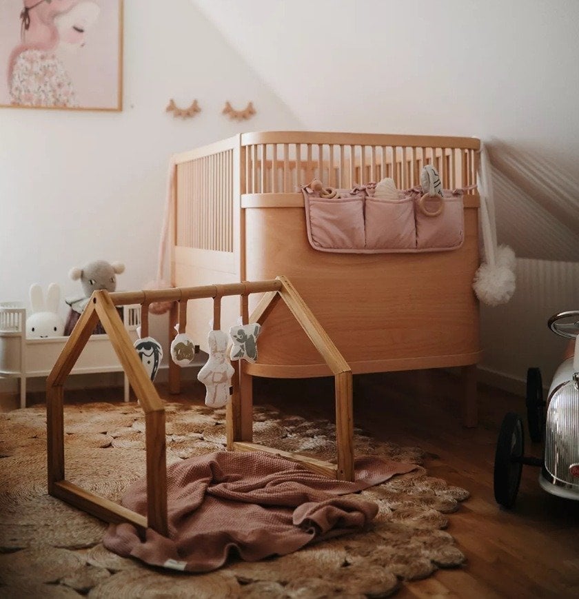 Drewniany stojak edukacyjny z zawieszonymi zabawkami dla niemowlaka w pokoju dziecięcym z łóżeczkiem i dywanem w kształcie kwiatu. Elodie Details House of Elodie Baby Gym Stojak edukacyjny