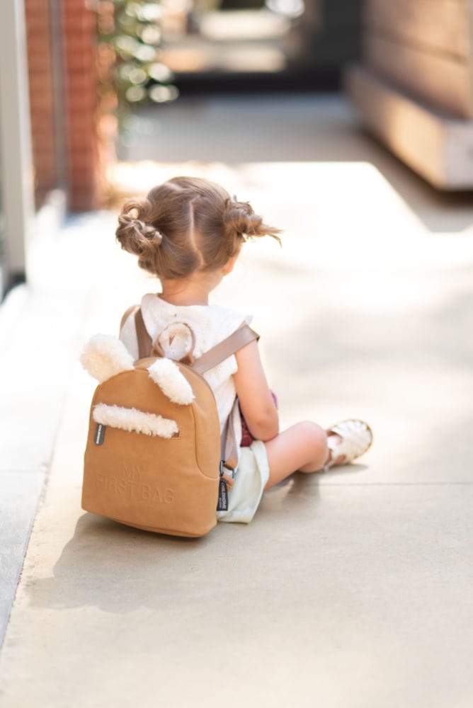 Dziecięcy plecak w kolorze jasnobrązowym z elementami przypominającymi futerko dla dziecka siedzącego na podłodze. Childhome Plecak dziecięcy My First Bag Suede-Look