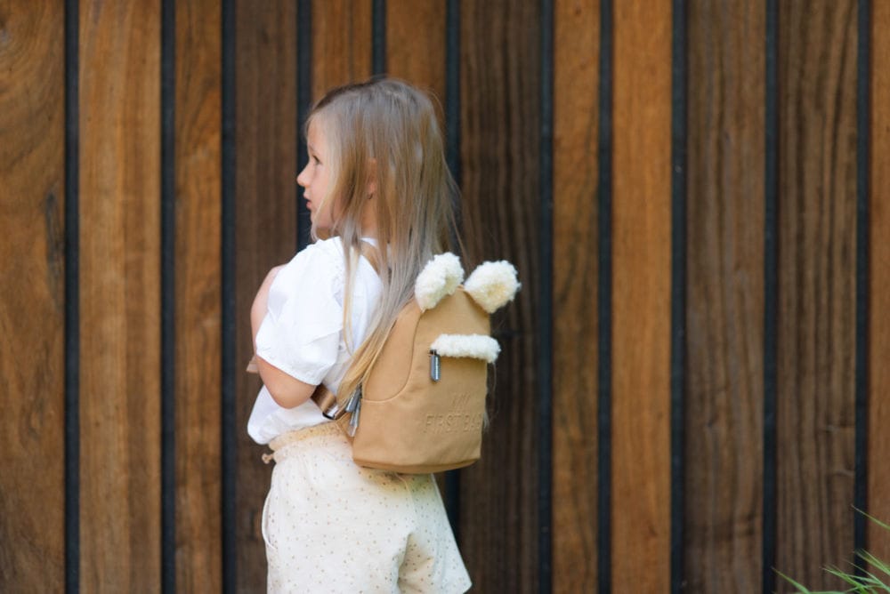 Dziecięcy plecak zamszowy w kolorze beżowym z uszami dla dziecka na spacerze na tle drewnianej ściany. Childhome Plecak dziecięcy My First Bag Suede-Look