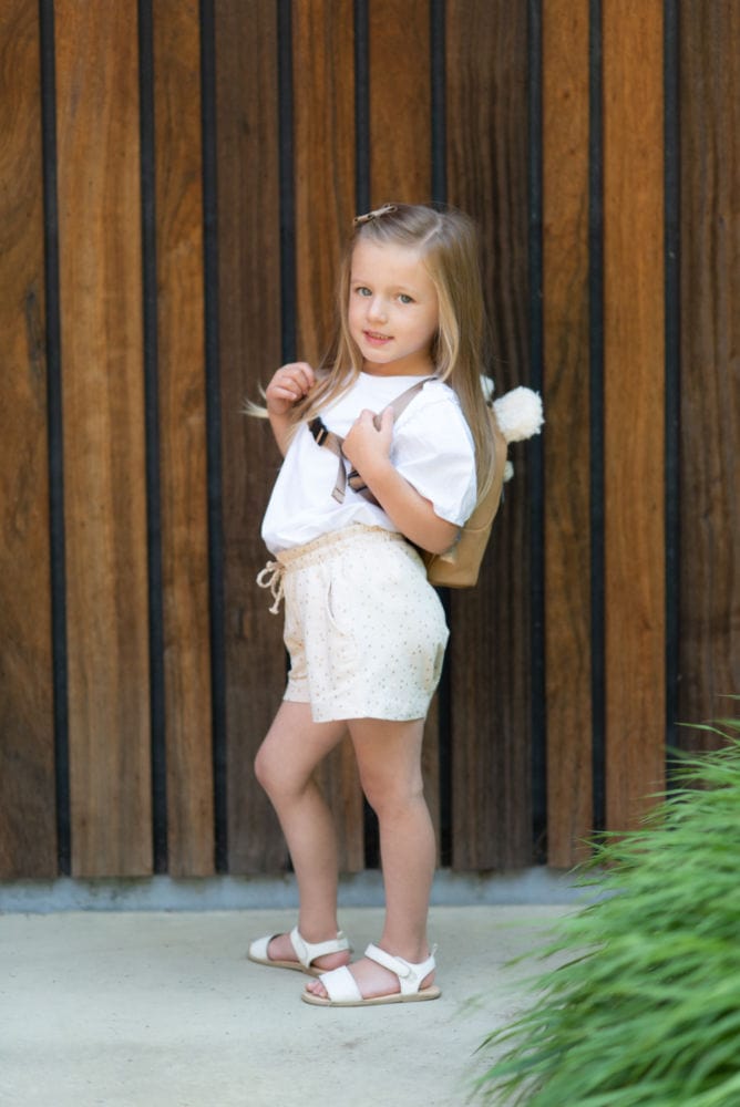 Dziecko z beżowym plecakiem dziecięcym z futerkiem na plecach ubrana w białą bluzkę i jasne spodenki na tle drewnianej ściany. Childhome Plecak dziecięcy My First Bag Suede-Look