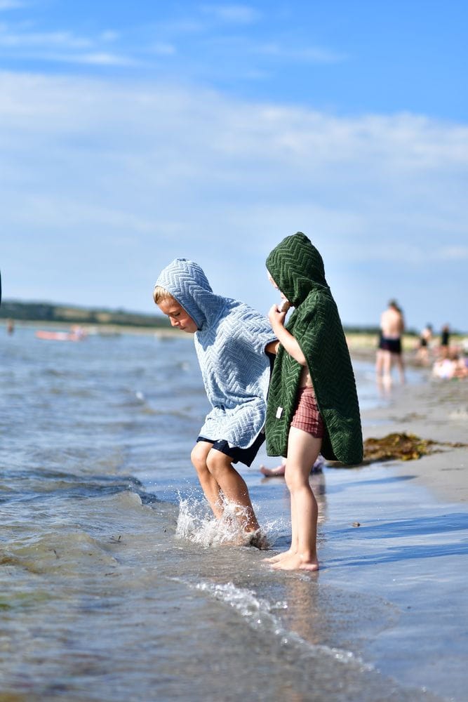 Dzieci na plaży w kąpielowych ponczach z kapturem w kolorze zielonym i niebieskim z wzorem zygzakowym. Filibabba Poncho Zigzag Dark Green