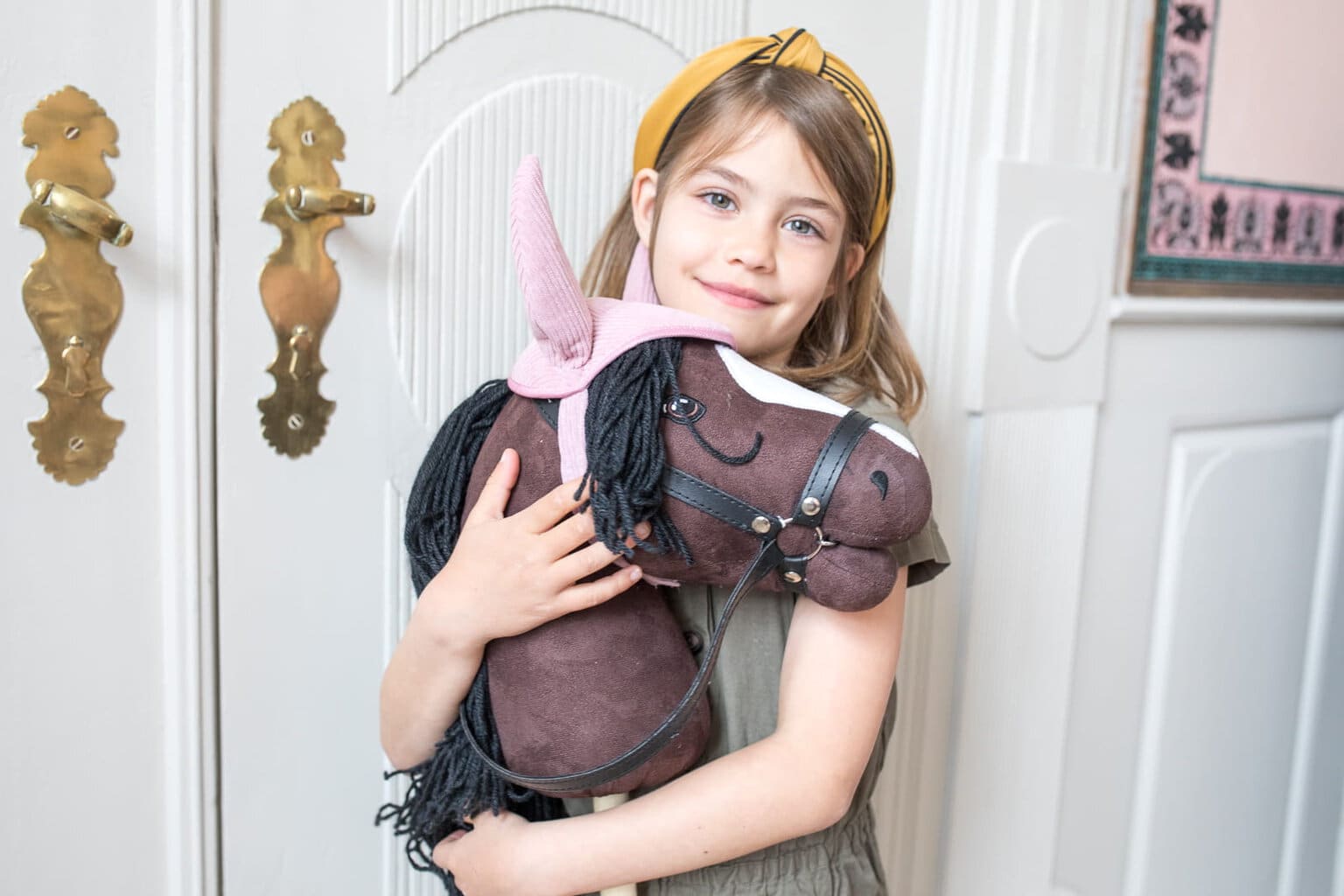 Dziewczynka trzyma brązowego konia na kiju z otwartą buzią i różowym rogiem jednorożca. byAstrup Hobby Horse Koń na kiju brązowy z otwartą buzią