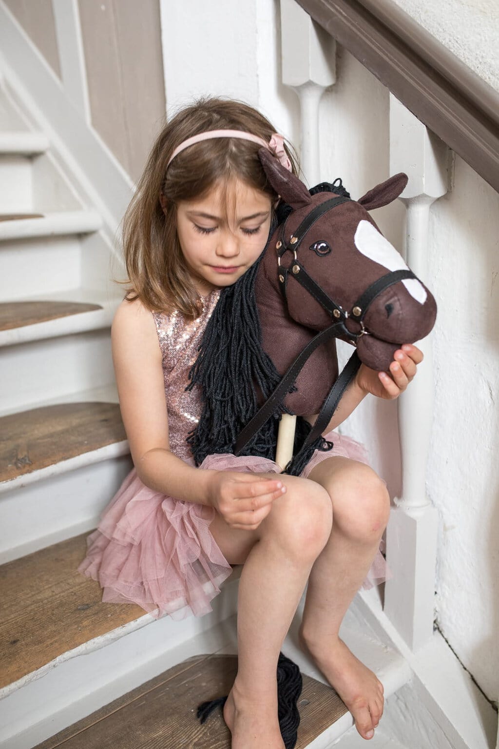Dziewczynka siedzi na schodach trzymając brązowego konia na kiju z otwartą buzią dla dziecka. byAstrup Hobby Horse Koń na kiju brązowy z otwartą buzią