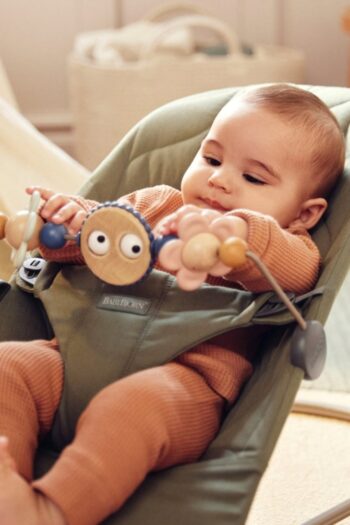 Ciemnozielony leżaczek dla niemowlaka z zabawkami na pałąku i dzieckiem w różowym ubranku. BABYBJORN Leżaczek BLISS Woven, Petal Quilt, Ciemnozielony