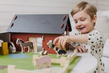 Drewniana czerwona farma w stylu szwedzkim z drewnianymi zwierzętami i dzieckiem bawiącym się na stole. MICKI Farma drewniana w stylu szwedzkim czerwona Micki