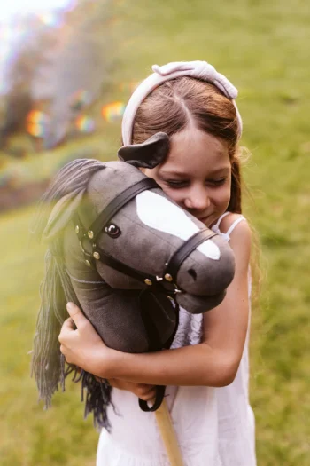Dziewczynka trzymająca szarego konia na kiju z otwartą buzią i czarną grzywą dla dziecka. byAstrup Hobby Horse szary koń na kiju z otwartą buzią i grzywą