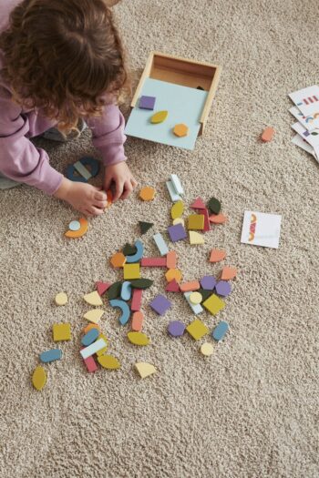 Dziecko układa drewnianą mozaikę z kolorowych elementów na dywanie dla dziecka. Kid’s Concept Drewniana układanka/puzzle-mozaika ZABAWKA ROKU 2023