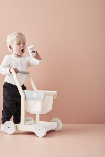 Drewniany wózek na zakupy dla dziecka z białym koszykiem i niebieskim panelem na tle beżowej ściany. Kid’s Concept Wózek na zakupy KID’S HUB