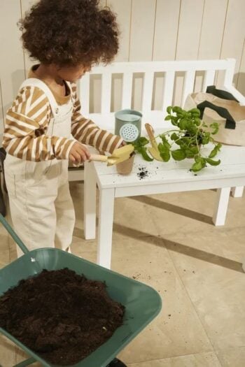 Dziecko bawiące się zestawem narzędzi ogrodowych z doniczką i rośliną na białej ławce oraz taczką z ziemią dla dziecka. Kid’s Concept Zestaw narzędzi ogrodowych KID’S HUB