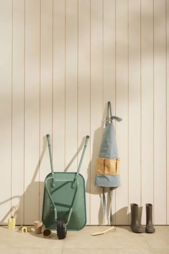 Fartuch ogrodnika dla dziecka w szaro-żółtym kolorze zawieszony na ścianie obok zielonej taczki i brązowych kaloszy. Kid’s Concept ​Fartuch ogrodnika KID’S HUB