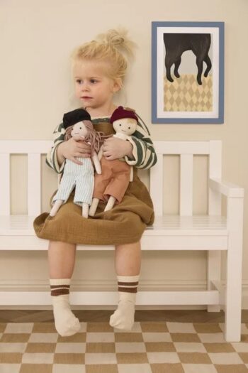 Dziewczynka siedzi na białej ławce trzyma dwie szmaciane lalki jedna w jasnych ubraniach druga w brązowych ubraniach. Kid’s Concept Lalka szmaciana Ingrid