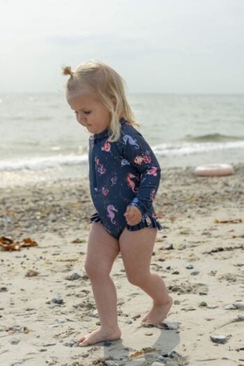Dziewczynka na plaży w granatowym kostiumie kąpielowym UV z motywem morskich zwierząt dla dziecka 3-4 lata. Filibabba Kostium UV Vilje (3-4 l) Rainbow Reef