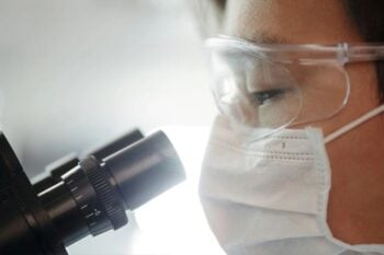 Kobieta w okularach ochronnych i maseczce medycznej patrząca w mikroskop