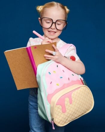 Dziewczynka z okularami nosi plecak dla dziecka w kształcie lodów z różowym i beżowym wzorem i pisze w notesie. Skip Hop Plecak dla Dziecka Lody Spark Style Little Kid