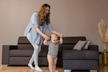 Kobieta pomaga niemowlakowi stawiać pierwsze kroki w salonie przy kanapie