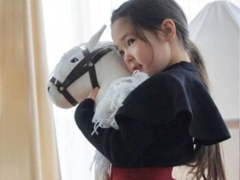Dziewczynka w czarnym sweterku i czerwonej spódnicy tiulowej Fig&Pear trzyma białego konia na patyku hobby horse, stoi obok białego namiotu dla dzieci