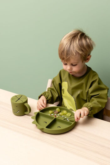 Zielony silikonowy trójdzielny talerz dla dziecka w kształcie dinozaura z przyssawką i dopasowany kubek z pokrywką na stoliku. Trixie Baby Dinozaur Silikonowy Talerz Trójdzielny z Przyssawką
