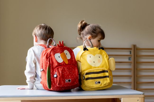 Dwa dzieci siedzące na stole z plecakami dziecięcymi w kształcie zwierząt czerwony z kurczakiem i żółty z pszczółką dla dziecka. Trixie Baby Pszczółka Plecak