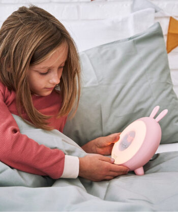 Dziewczynka w łóżku włącza różową lampkę budzik w kształcie królika dla dziecka. Rabbit&Friends Królik Lampka Budząca Światłem Różowa