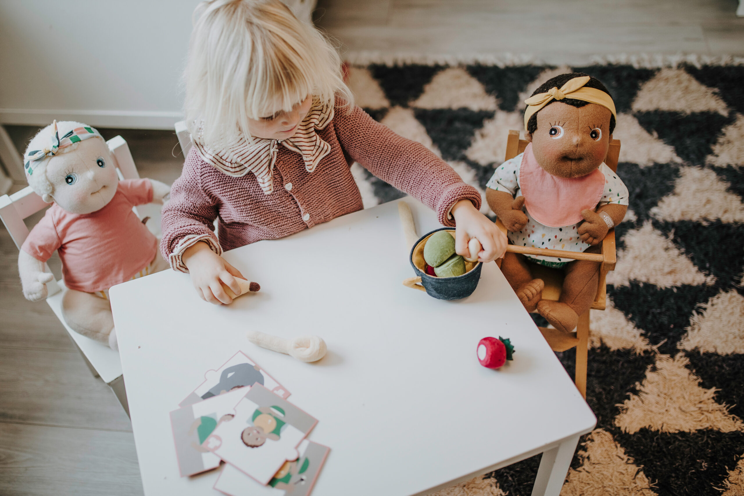 Dziecko bawiące się z dwiema szmacianymi lalkami siedzącymi przy białym stoliku na dywanie z geometrycznym wzorem. Rubens Barn Anatomiczna lalka sensoryczna Carl Baby