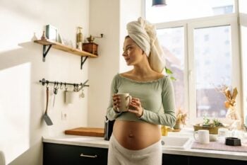 Kobieta w ciąży w kuchni trzymająca filiżankę kawy z ręcznikiem na głowie
