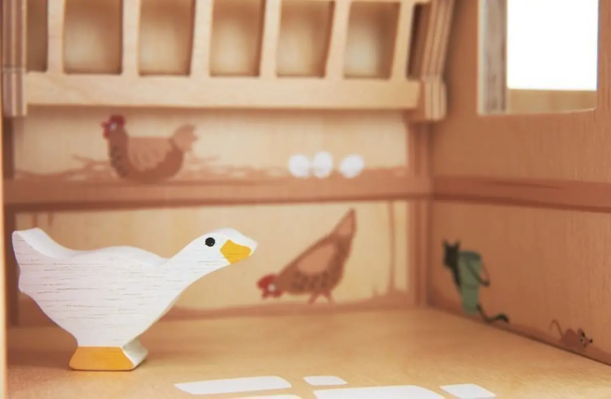 Drewniana biała figurka gęsi z żółtym dziobem i stopami na tle drewnianej zabawkowej farmy. Tender Leaf Toys Drewniane figurki do zabawy farma z zwierzątkami