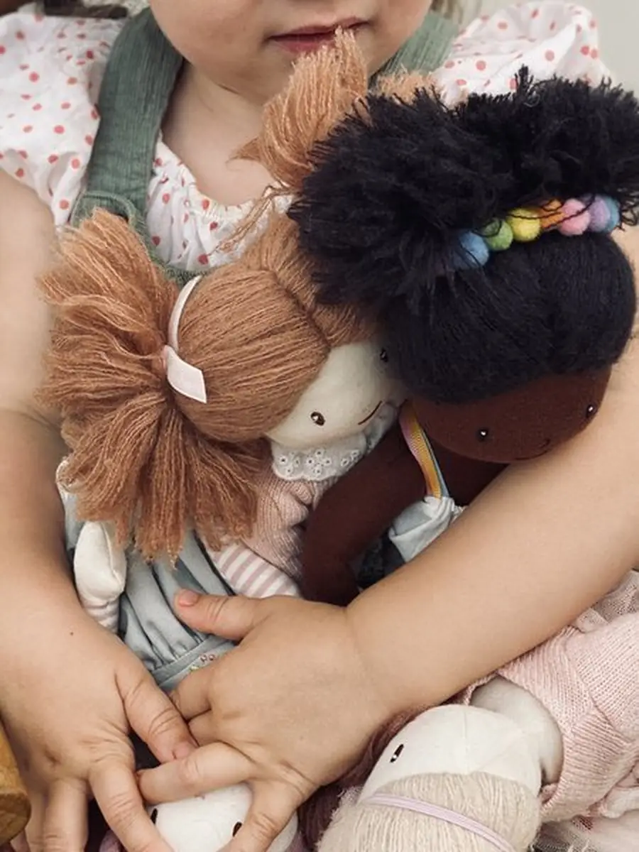 Dziecko trzyma dwie szmaciane lalki jedna z rudymi włosami druga z czarnymi kręconymi włosami dla dziecka. ThreadBear Design Lalka Esme