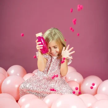 Dziewczynka bawiąca się różową butelką sensoryczną siedząc wśród różowych balonów dla małego odkrywcy. Petit Boum Butelka sensoryczna FLOAT różowa