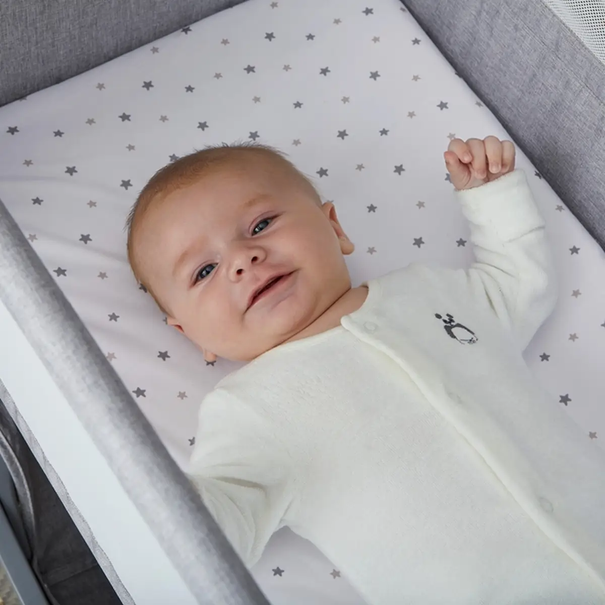 Białe prześcieradło z szarymi gwiazdkami na łóżeczku dziecięcym z niemowlakiem w białym pajacyku. Shnuggle Prześcieradło 80x40cm Air Lite Crib 2szt Stars