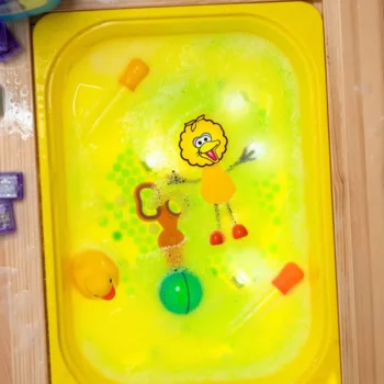 Żółta plastikowa taca z pianką do kąpieli i zabawkami w tym żółty ptak i dwie świecące kostki sensoryczne dla dziecka do kąpieli. Glo Pals Postać do kąpieli Wielki Ptak zestaw z dwiema kostkami sensorycznymi świecącymi w wodzie