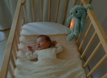 Drewniane łóżeczko dziecięce z niemowlakiem i szumiącą lampką nocną królik w kolorze szałwiowym zawieszoną na barierce. Zazu RO Szumiąca Lampka Nocna Królik Sage