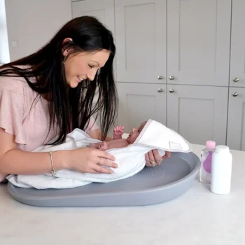 Szara piankowa mata do przewijania na komodę z niemowlakiem owiniętym w biały ręcznik dla mamy. Shnuggle Mata do Przewijania z Pianki Squishy Grey
