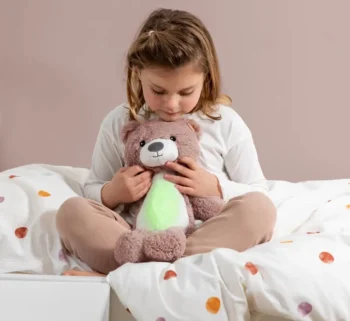 Dziewczynka siedzi na łóżku trzyma różowego pluszowego misia szumiącą przytulankę dla dziecka. Zazu BRODY Trener Snu i Szumiąca Przytulanka Miś Pink