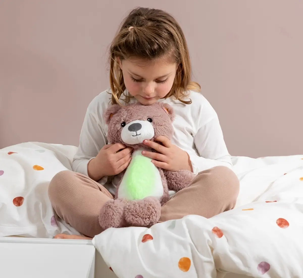 Dziewczynka siedzi na łóżku trzyma różowego pluszowego misia szumiącą przytulankę dla dziecka. Zazu BRODY Trener Snu i Szumiąca Przytulanka Miś Pink