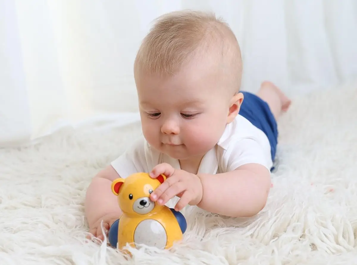 Niemowlę bawiące się żółtym wańka wstańka w kształcie misia na jasnym dywanie. Tolo Bio Miś Roly Poly 6m+