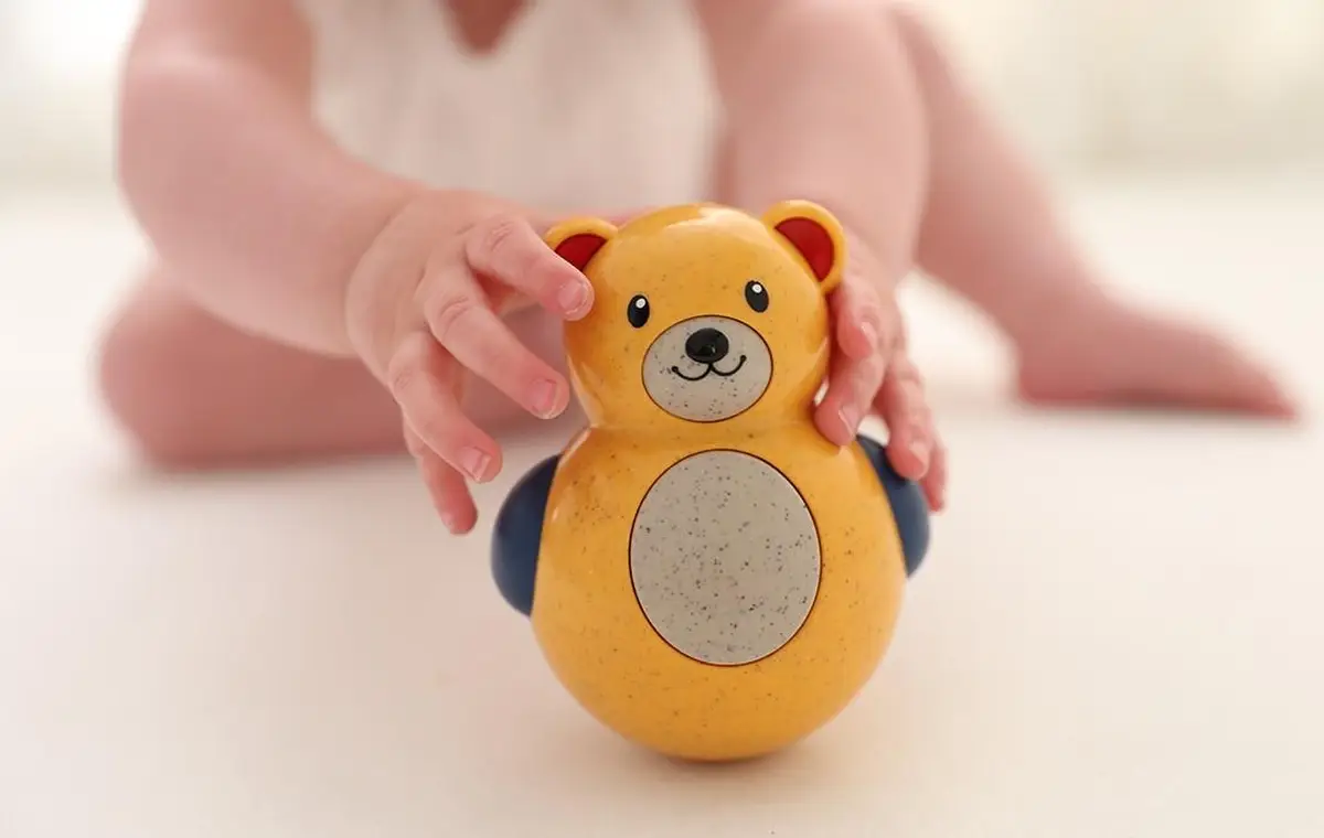 Żółty wańka wstańka miś z niebieskimi łapkami trzymany przez dziecko dla niemowlaka. Tolo Bio Miś Roly Poly 6m+