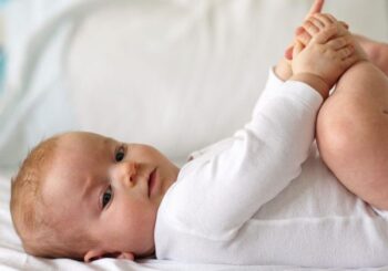 Niemowlę w białej piżamce leżące na plecach trzymające stopy rękami dla niemowlaka 7 miesięcy