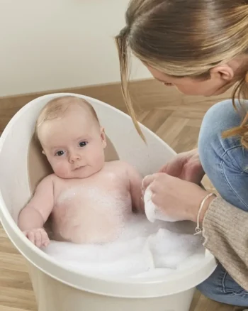 Wanienka dla niemowlaka w kolorze beżowym z odpływem z pianką do kąpieli i dzieckiem w środku oraz osobą myjącą dziecko. Shnuggle ECO Wanienka dla Niemowlaka z Odpływem Baby