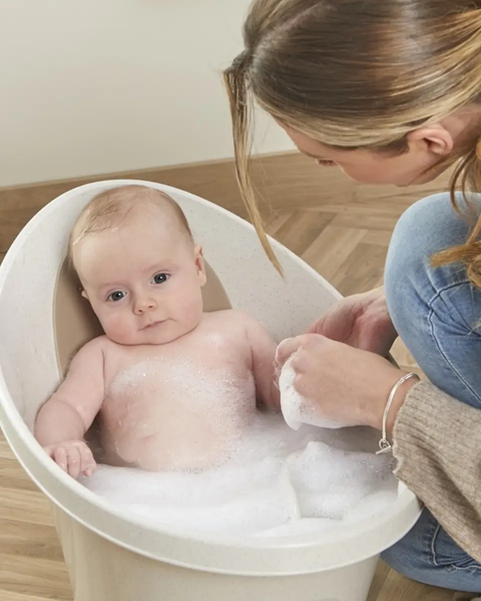 Wanienka dla niemowlaka w kolorze beżowym z odpływem z pianką do kąpieli i dzieckiem w środku oraz osobą myjącą dziecko. Shnuggle ECO Wanienka dla Niemowlaka z Odpływem Baby