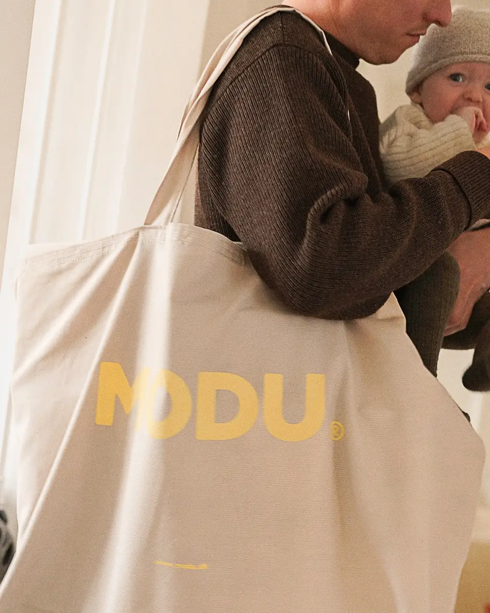 Beżowa torba transportowa z żółtym napisem modu na ramieniu osoby trzymającej niemowlę. MODU torba transportowa Sand Grey / Honey Yellow