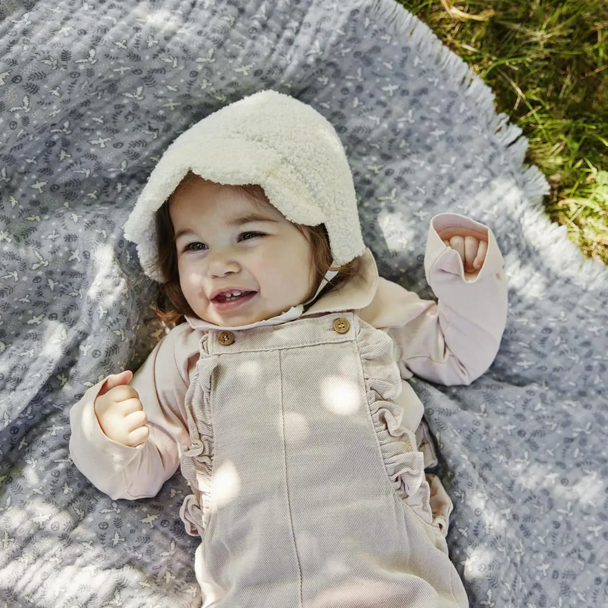 Niemowlę w białej czapce bouclé leżące na szarym kocyku z wzorem dla niemowlaka. Elodie Details Czapka Baby Bonnet White Bouclé 0-3 m-cy