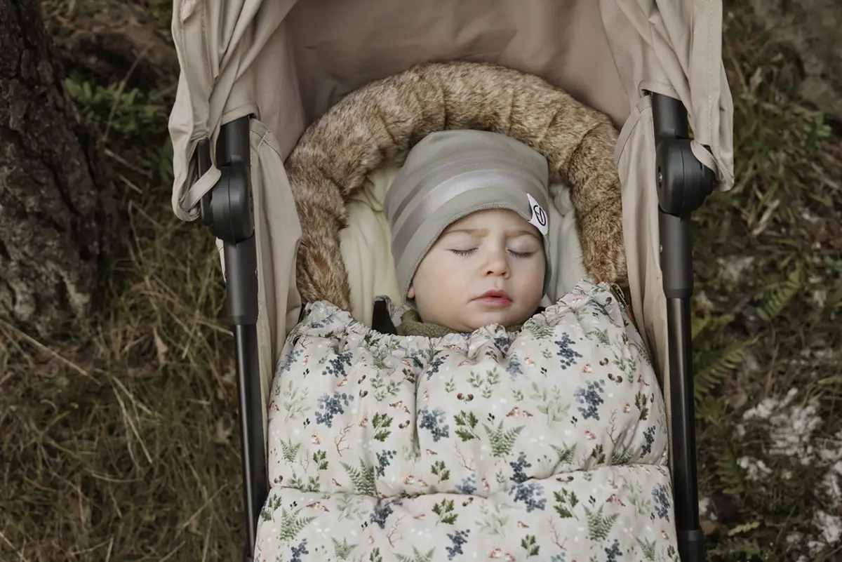 Niemowlę w szarej zimowej czapce w paski leżące w wózku z kwiatowym śpiworkiem na zewnątrz. Elodie Details Czapka Hazy Jade 0-6 m-cy
