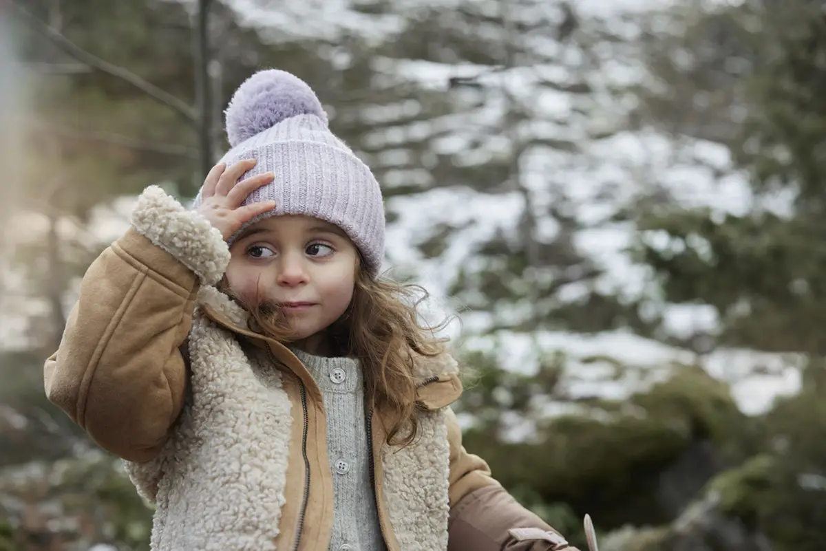 Dziewczynka w jasnofioletowej wełnianej czapce z pomponem zimowej dla dziecka na tle zimowego lasu. Elodie Details Czapka Wełniana Lavender Love 0-6 m-cy