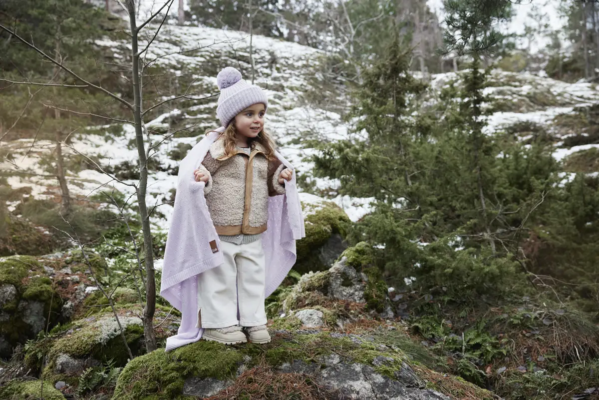 Dziewczynka w jasnofioletowej wełnianej czapce zimowej z pomponem na tle lasu z resztkami śniegu dla dziecka. Elodie Details Czapka Wełniana Lavender Love 0-6 m-cy