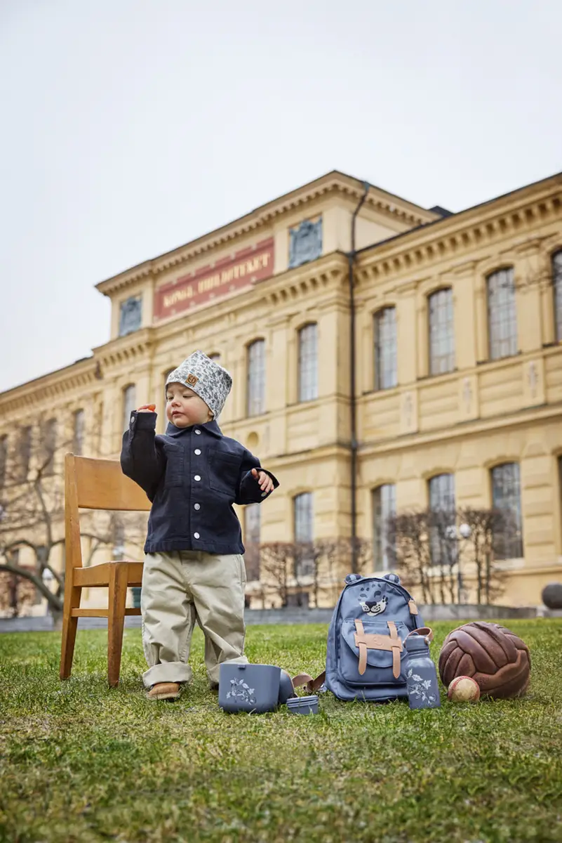 Butelka na wodę z nadrukiem roślinnym w szarym kolorze dla dziecka obok plecaka i pojemnika na przekąski na trawie. Elodie Details Butelka na wodę Garden Leo