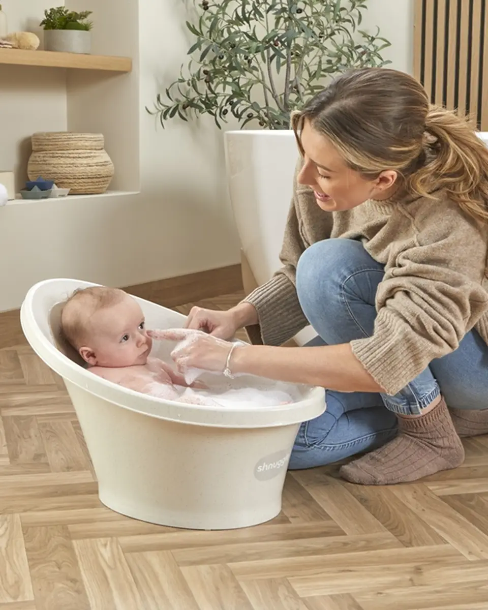 Wanienka dla niemowlaka w kolorze ecru z odpływem dziecko w kąpieli kobieta w swetrze i dżinsach siedzi obok na podłodze drewnianej. Shnuggle ECO Wanienka dla Niemowlaka z Odpływem Baby