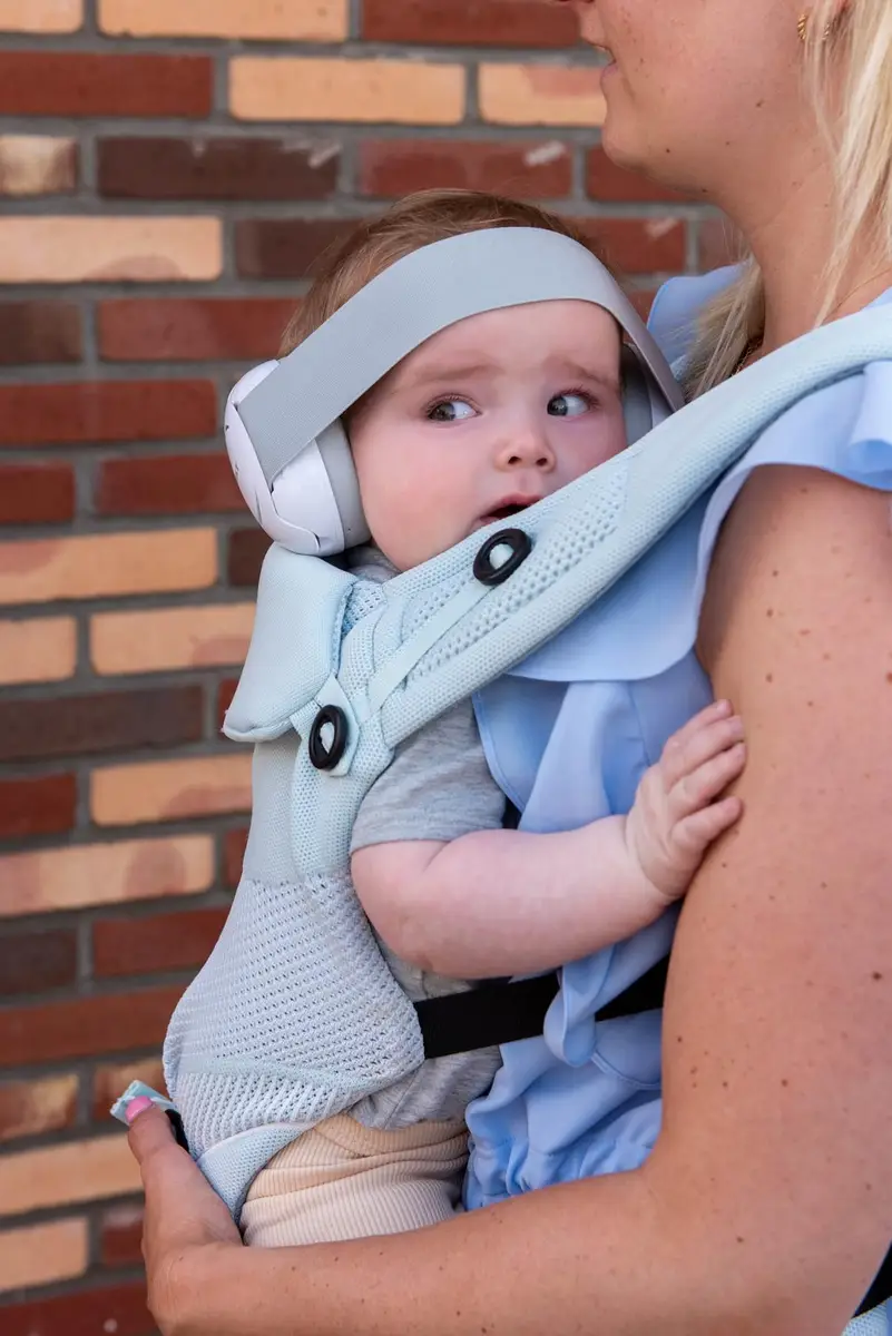 Niemowlę w jasnoszarych słuchawkach ochronnych na uszach trzymane przez kobietę w nosidełku dla dziecka na tle ceglanej ściany. Dooky Słuchawki ochronne Baby Earmuff white 0-3 l